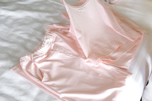 A blush pink cotton pajama set folded on white bedding, showing the lace trimmed waistband, satin bow and mother of pearl buttons