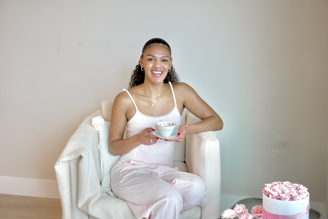 A woman sitting in a cream armchair in a blush cotton pajama set, holding a teacup and saucer, pink roses on a table beside her