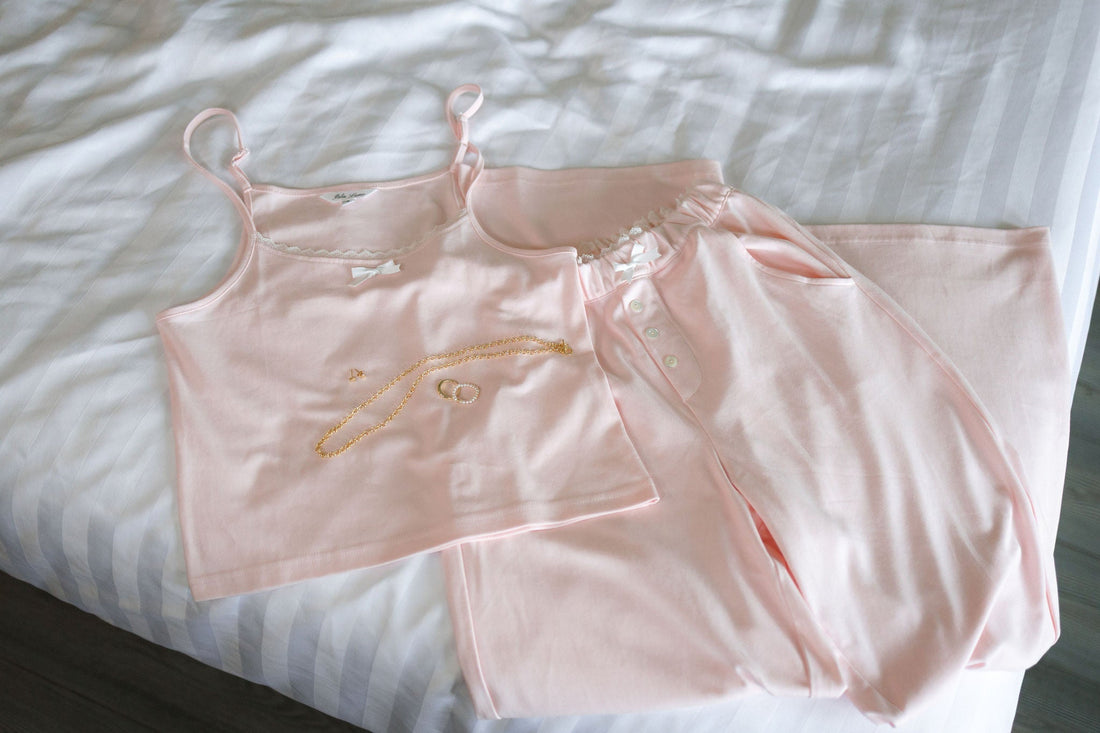 A blush 100% cotton pajama set laid flat on white striped bedding with a gold necklace and rings