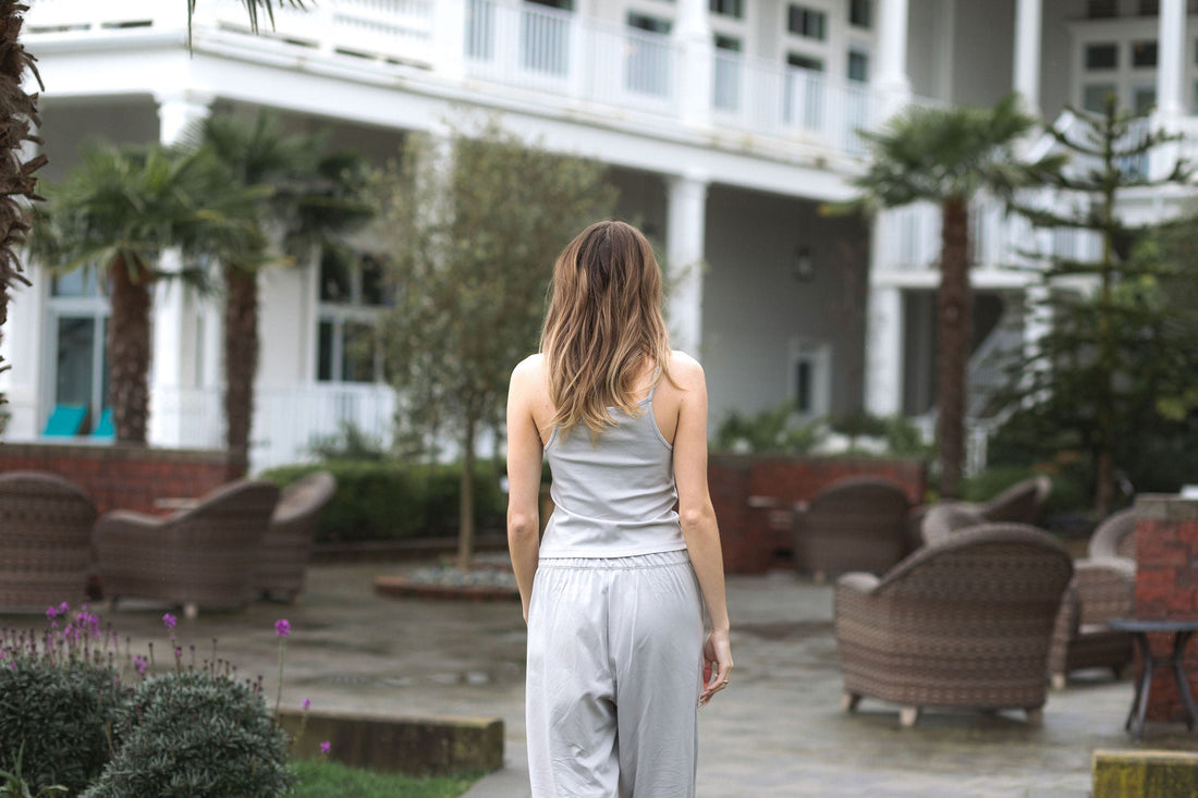 A woman walking through a hotel courtyard in a Stone grey cotton pajama set, seen from behind, with palm trees and wicker chairs around her