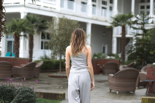 A woman walking through a hotel courtyard in a Stone grey cotton pajama set, seen from behind, with palm trees and wicker chairs around her