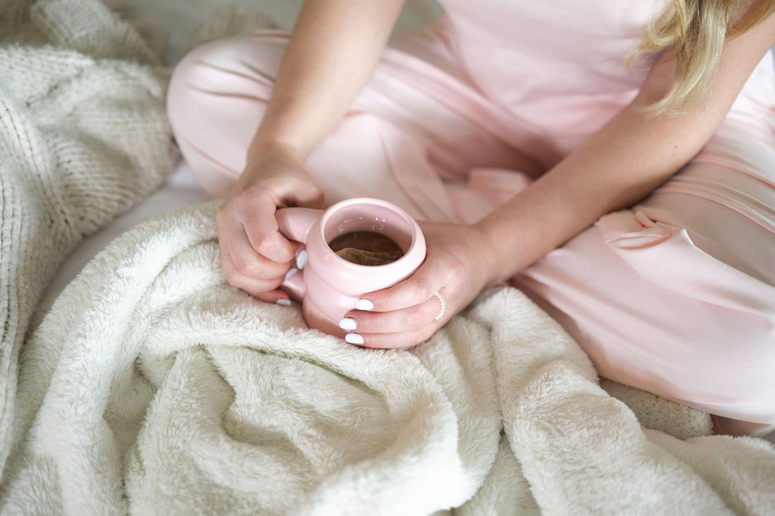Hands holding a pink mug of tea in a lap, wearing blush cotton pajama pants under a cream blanket