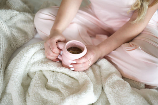Hands holding a pink mug of tea in a lap, wearing blush cotton pajama pants under a cream blanket