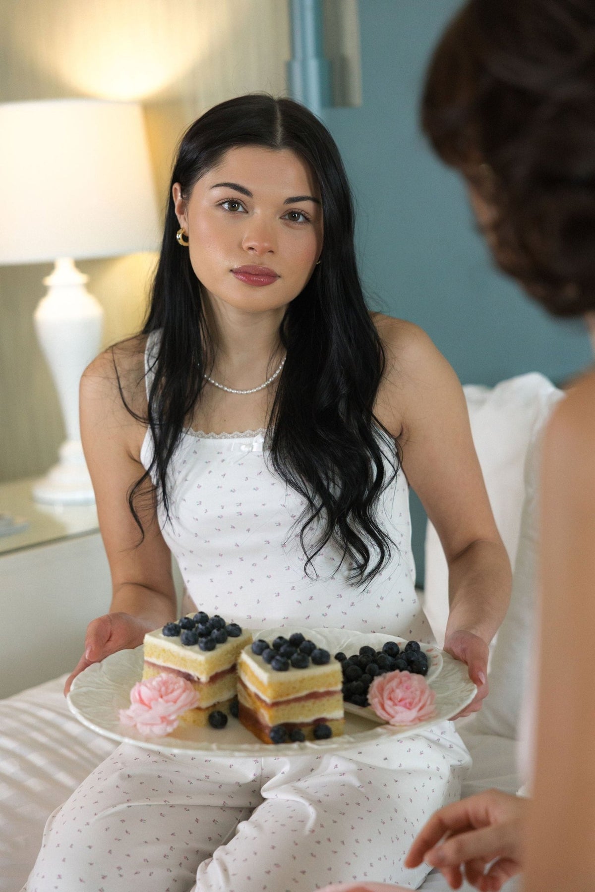 The Love Letter cotton pajama top worn with the matching floral pants, seated on a bed holding a plate of berry cakes